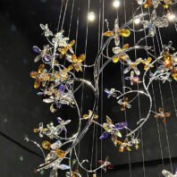 A stunning large chandelier exemplifies innovative custom lighting design in a hotel interior. This big chandelier features intricate, jewel-toned crystal flowers suspended on delicate silver wires and branches. The hotel chandelier creates a magical floating garden effect, with amber, purple, and clear crystal blossoms seemingly in flight. Warm LED lights illuminate the crystals, causing them to sparkle brilliantly against a dark background. This custom lighting masterpiece transforms the space into an enchanted forest, beautifully merging nature-inspired artistry with contemporary luxury, perfect for creating a mesmerizing focal point in a modern hotel lobby or dining area.