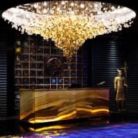 A spectacular large chandelier illuminates a hotel lobby, exemplifying exquisite custom lighting design. This big chandelier features a golden cascade of crystal-like elements, transitioning from clear to amber, creating a breathtaking focal point. The hotel chandelier hovers above a striking backlit onyx reception desk. Behind, an intricate wall display showcases ornate designs, while a golden statue stands guard. This custom lighting masterpiece transforms the space into a luxurious, awe-inspiring entrance, perfectly balancing modern opulence with artistic flair.