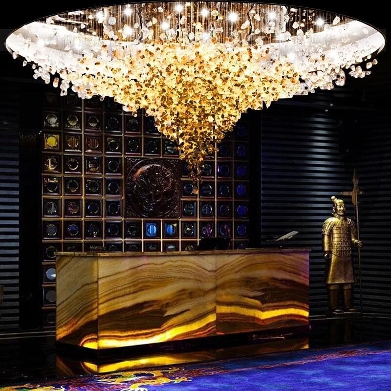 A spectacular large chandelier illuminates a hotel lobby, exemplifying exquisite custom lighting design. This big chandelier features a golden cascade of crystal-like elements, transitioning from clear to amber, creating a breathtaking focal point. The hotel chandelier hovers above a striking backlit onyx reception desk. Behind, an intricate wall display showcases ornate designs, while a golden statue stands guard. This custom lighting masterpiece transforms the space into a luxurious, awe-inspiring entrance, perfectly balancing modern opulence with artistic flair.