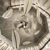 An innovative custom lighting installation featuring multiple leaf-shaped light fixtures suspended within a spiral staircase. The chandelier consists of numerous illuminated white structures resembling stylized leaves or feathers, each with a golden spine. These leaf-like elements are suspended at various heights and angles, creating a dynamic, floating effect. The installation is viewed from below, showcasing the circular arrangement that follows the curve of the staircase. Thin cables support each light fixture, adding to the illusion of weightlessness. The spiral staircase's railing is visible around the perimeter, framing the ethereal lighting display. This bespoke lighting design transforms the stairwell into a mesmerizing, nature-inspired art piece, demonstrating how custom lighting can elevate architectural spaces with artistic and functional elements.