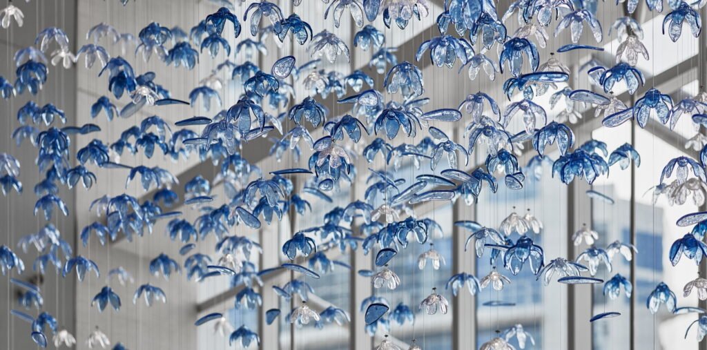 A stunning custom lighting installation featuring a large chandelier composed of hundreds of delicate, blue and clear glass flower-shaped elements. This big hotel chandelier creates an illusion of an underwater garden, with translucent petals suspended on thin wires at varying heights. The blue-tinted glass flowers dominate, interspersed with clear ones, giving depth and dimension to the piece. Sunlight streams through nearby windows, interacting with the chandelier to cast intricate shadows and reflections. Some petals appear elongated, adding to the fluid, aquatic feel. This bespoke lighting design transforms the hotel space into a mesmerizing, ocean-inspired wonderland, showcasing how custom lighting can create an immersive, artistic environment that captivates guests and elevates the hotel's unique aesthetic appeal.