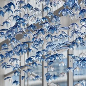 A stunning custom lighting installation featuring a large chandelier composed of hundreds of delicate, blue and clear glass flower-shaped elements. This big hotel chandelier creates an illusion of an underwater garden, with translucent petals suspended on thin wires at varying heights. The blue-tinted glass flowers dominate, interspersed with clear ones, giving depth and dimension to the piece. Sunlight streams through nearby windows, interacting with the chandelier to cast intricate shadows and reflections. Some petals appear elongated, adding to the fluid, aquatic feel. This bespoke lighting design transforms the hotel space into a mesmerizing, ocean-inspired wonderland, showcasing how custom lighting can create an immersive, artistic environment that captivates guests and elevates the hotel's unique aesthetic appeal.