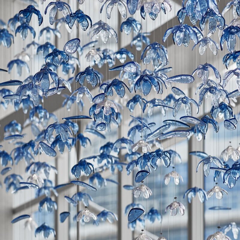 A stunning custom lighting installation featuring a large chandelier composed of hundreds of delicate, blue and clear glass flower-shaped elements. This big hotel chandelier creates an illusion of an underwater garden, with translucent petals suspended on thin wires at varying heights. The blue-tinted glass flowers dominate, interspersed with clear ones, giving depth and dimension to the piece. Sunlight streams through nearby windows, interacting with the chandelier to cast intricate shadows and reflections. Some petals appear elongated, adding to the fluid, aquatic feel. This bespoke lighting design transforms the hotel space into a mesmerizing, ocean-inspired wonderland, showcasing how custom lighting can create an immersive, artistic environment that captivates guests and elevates the hotel's unique aesthetic appeal.