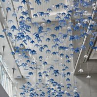 A breathtaking custom lighting installation features a large chandelier composed of hundreds of translucent blue and clear glass petal-shaped elements. This big hotel chandelier creates an illusion of an underwater garden, with delicate petals suspended on thin wires at varying heights. The blue-tinted glass petals dominate, interspersed with clear ones, giving depth and dimension to the piece. Natural light from nearby windows interacts with the chandelier, casting intricate shadows and reflections on the white walls and ceiling. This bespoke lighting design transforms the hotel's high-ceilinged space into a mesmerizing, aquatic-inspired wonderland, showcasing how custom lighting can create an immersive, artistic environment that captivates guests and elevates the hotel's unique aesthetic appeal.