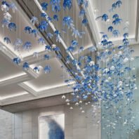 A stunning custom lighting installation adorns the ceiling of a luxurious hotel lobby. This large chandelier features hundreds of delicate blue and clear glass flower-shaped elements suspended at varying heights, creating a cascading effect. The big hotel chandelier spans the entire ornate ceiling, with blue blossoms dominating and clear ones interspersed throughout. Soft, recessed lighting in the ceiling coves enhances the chandelier's ethereal glow. The installation complements the lobby's modern design, featuring sleek marble walls and a hint of blue artwork. This bespoke lighting transforms the space into an enchanting underwater garden, demonstrating how custom lighting can create an immersive, artistic environment that captivates guests upon entry.