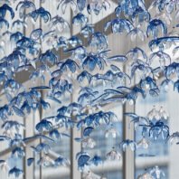 A stunning custom lighting installation featuring a large chandelier composed of hundreds of delicate, blue and clear glass flower-shaped elements. This big hotel chandelier creates an illusion of an underwater garden, with translucent petals suspended on thin wires at varying heights. The blue-tinted glass flowers catch and refract light, casting intricate shadows and giving the impression of gentle movement. This bespoke lighting design transforms the hotel space into a mesmerizing, aquatic-inspired wonderland, demonstrating how custom lighting can create an immersive, artistic environment that captivates guests and elevates the hotel's aesthetic appeal.