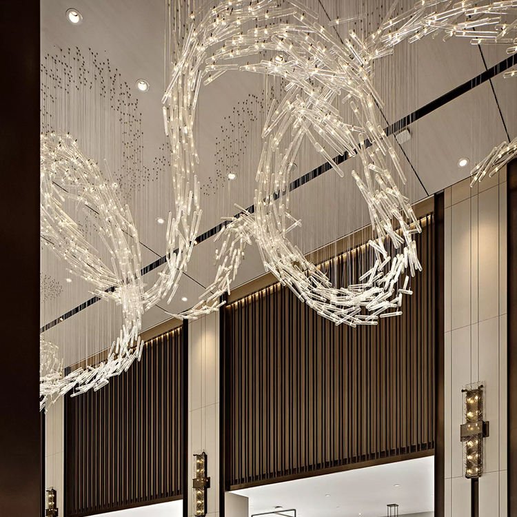 A mesmerizing custom lighting installation adorns the ceiling of a luxurious hotel lobby. This large chandelier features intertwining ribbons of illuminated glass tubes, creating fluid, wave-like forms that sweep across the space. The big chandelier's dynamic design mimics the graceful movement of wind or water, its warm light contrasting beautifully with the room's neutral tones. Delicate strands of tiny lights cascade from the ceiling, adding depth and sparkle. The hotel's interior combines sleek modern elements with elegant vertical paneling and subtle wall sconces. This bespoke hotel chandelier transforms the lobby into an enchanting, otherworldly environment, showcasing how custom lighting can create an unforgettable first impression for guests.