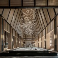 A breathtaking custom lighting installation dominates a luxurious hotel atrium. This large chandelier features an intricate, abstract design of illuminated white rods intersecting at various angles, resembling a frozen starburst or lightning storm. The big chandelier spans the vaulted ceiling, creating a dramatic focal point. The hotel's elegant corridor showcases modern architecture with sleek lines, warm wood tones, and vertical lighting fixtures along the walls. At the far end, a illuminated tree sculpture adds a touch of nature. A reflective pool in the foreground mirrors the chandelier, enhancing its impact. This bespoke hotel chandelier transforms the space into a captivating blend of contemporary design and artistic expression.