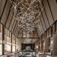 A stunning custom lighting installation dominates the atrium of a luxurious hotel. This large chandelier features an intricate, abstract design of illuminated white rods intersecting at various angles, resembling a frozen starburst or lightning storm. The big chandelier spans the entire vaulted ceiling, creating a dramatic focal point. Below, the hotel's elegant lobby showcases modern furnishings, warm wood tones, and floor-to-ceiling windows. At the far end, a illuminated tree sculpture adds a touch of nature. This bespoke hotel chandelier transforms the space into a captivating blend of contemporary design and artistic expression, exemplifying how custom lighting can create an unforgettable guest experience.