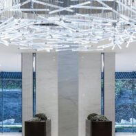 A striking large chandelier dominates the ceiling of a contemporary hotel lobby. This custom lighting fixture features an intricate geometric design of intersecting white LED tubes, creating a web-like pattern. The big chandelier spans the entire ceiling, casting a bright, even light across the space. Below, sleek marble columns frame floor-to-ceiling windows, offering views of a serene outdoor landscape. Two modern planters with lush greenery flank the entrance, complementing the hotel chandelier's avant-garde design and enhancing the lobby's sophisticated ambiance.