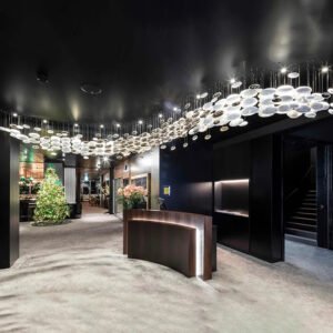 A large custom lighting chandelier dominates a luxurious hotel lobby. This big chandelier features hundreds of suspended disc-shaped elements, creating a flowing, wave-like pattern across the dark ceiling. The hotel chandelier blends illuminated white and reflective metallic surfaces, resembling a celestial stream. Below, a curved reception desk with subtle lighting stands near a festively decorated Christmas tree. The lobby's design combines contemporary elements with warm accents, showcasing how custom lighting can transform a space into an inviting, memorable environment.