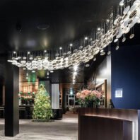 A stunning modern hotel lobby featuring an eye-catching custom lighting installation. The centerpiece is a large, undulating chandelier composed of numerous disc-shaped elements suspended at varying heights. These discs, a mix of illuminated white and reflective metallic surfaces, create a flowing, wave-like pattern across the dark ceiling, reminiscent of a celestial stream or galaxy. The lobby's design is a sophisticated blend of contemporary and warm elements. A sleek reception desk with subtle under-lighting is visible in the foreground, adorned with a vibrant arrangement of pink flowers. To the left, a festively decorated Christmas tree adds a touch of seasonal charm and warmth to the space. The walls feature a combination of dark panels and wood accents, creating a rich, luxurious atmosphere. The floor appears to be a polished concrete or stone, reflecting some of the overhead lighting and adding to the space's sense of openness. In the background, glimpses of a bar area can be seen, suggesting this lobby may lead to additional amenities such as a restaurant or lounge. The overall effect is one of modern elegance, with the innovative lighting design serving as both a functional light source and a captivating art installation, transforming the lobby into a memorable and inviting space for hotel guests.