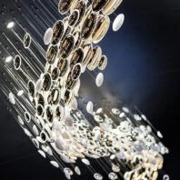 A captivating close-up of a contemporary chandelier installation featuring a multitude of suspended disc-shaped elements. The design creates a flowing, wave-like pattern against a dark background. The discs vary in size and material, including highly reflective gold or bronze surfaces, transparent elements, and opaque white discs. Each piece is suspended on thin, nearly invisible wires at different heights, creating a three-dimensional, cascading effect. The arrangement transitions from larger, more densely packed discs at the top to smaller, more dispersed elements towards the bottom, giving the impression of a celestial shower or a dynamic, abstract waterfall. Light sources integrated within the installation cause the metallic surfaces to gleam and create intriguing reflections and shadows. This avant-garde lighting fixture transforms the space into a mesmerizing, luxurious environment, perfect for upscale venues such as high-end restaurants, hotel lobbies, or modern art galleries.