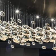 A striking modern chandelier featuring a multitude of suspended disc-shaped elements creates a mesmerizing display against a dark ceiling. The installation consists of numerous metallic and transparent discs of varying sizes, suspended at different heights on thin wires. The discs appear to be a mix of highly reflective surfaces, likely chrome or polished metal, and clear glass or acrylic. The arrangement forms a dynamic, wave-like pattern that seems to flow across the ceiling, reminiscent of planetary orbits or a celestial stream. Light sources embedded within or above the installation cause the discs to gleam and sparkle, creating an interplay of light and shadow. This avant-garde lighting fixture transforms the space into a futuristic, luxurious environment, perfect for high-end venues such as upscale restaurants, hotel lobbies, or contemporary art galleries. The overall effect is one of suspended motion, lending a sense of elegance and intrigue to the room.