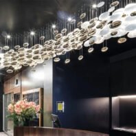 An impressive custom lighting installation adorns the ceiling of a modern reception area. The chandelier features a dynamic arrangement of suspended disc-shaped elements in various sizes, creating a flowing, wave-like pattern across the ceiling. The discs appear to be a mix of illuminated white circles and reflective gold surfaces, suspended at different heights on thin wires. This creates a mesmerizing, three-dimensional effect reminiscent of a celestial stream or abstract constellation. The lighting design contrasts beautifully with the dark ceiling, casting a warm, inviting glow over the space. Below, a reception desk is visible, adorned with a floral arrangement of pink flowers. The walls feature a mix of materials, including what appears to be wood paneling and dark-colored surfaces, complementing the modern, luxurious aesthetic of the chandelier. This striking lighting fixture serves as a focal point, transforming the reception area into an elegant and visually captivating space.