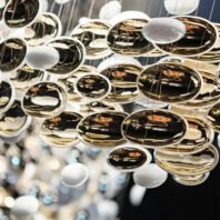 A close-up view of an avant-garde chandelier featuring an array of suspended glass or acrylic discs in various sizes. The discs are a mix of transparent, reflective gold, and opaque white elements, creating a dynamic interplay of light and reflection. Some of the larger discs appear to have a concave shape, enhancing their reflective properties. The arrangement creates a sense of depth and movement, with the discs seemingly floating at different levels. The reflective surfaces capture and distort the surrounding environment, including what appears to be warm lighting from the room. This intricate lighting fixture exemplifies contemporary luxury design, suitable for high-end spaces such as upscale restaurants, hotel lobbies, or exclusive retail environments. The overall effect is one of sophisticated elegance with a touch of cosmic inspiration, reminiscent of planets or celestial bodies suspended in space.