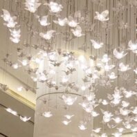 A close-up view of an intricate custom lighting installation featuring a multitude of illuminated butterfly-shaped elements. The chandelier consists of numerous delicate, translucent butterflies suspended at varying heights on thin wires, creating a three-dimensional, floating effect. Each butterfly glows softly, emitting a gentle, warm light that gives the installation an ethereal quality. The butterflies appear to be made of a frosted or crystal-like material, allowing the light to diffuse beautifully through their forms. In the background, a cylindrical structural column can be partially seen, hinting at the grandeur of the space. This detailed view showcases the exquisite craftsmanship of the lighting design, which transforms the area into a magical, dream-like environment reminiscent of a fluttering swarm of luminous butterflies.