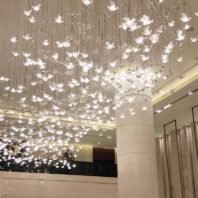 An extraordinary custom lighting installation dominates the ceiling of an elegant hotel atrium. The chandelier features hundreds of illuminated butterfly-shaped elements suspended at various heights on thin wires, creating a mesmerizing, three-dimensional effect. Each delicate butterfly glows softly, emitting a warm, ethereal light that bathes the space in a gentle luminescence. The installation spans the entire visible ceiling, giving the illusion of a vast swarm of glowing butterflies frozen in mid-flight. This enchanting lighting design is complemented by the atrium's modern architecture, including a tall cylindrical column and a visible mezzanine level with glass balustrades. The combination of the whimsical, nature-inspired lighting with the sleek, contemporary interior transforms the atrium into a magical and welcoming space, creating a memorable and awe-inspiring atmosphere for hotel guests.