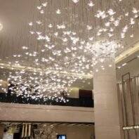 A stunning custom lighting installation adorns the ceiling of a spacious hotel atrium. The chandelier features hundreds of illuminated butterfly-shaped elements suspended at varying heights, creating a magical, floating effect. Each delicate butterfly glows softly, emitting a warm, diffused light that fills the space with a gentle radiance. The installation spans the entire ceiling, giving the impression of a vast swarm of luminous butterflies in mid-flight. This enchanting lighting design is complemented by the atrium's modern architecture, including tall cylindrical columns and a visible mezzanine level. The lower level of the atrium is partially visible, showcasing additional decorative elements and what appears to be a digital display. The combination of the whimsical, nature-inspired lighting with the sleek, contemporary interior transforms the atrium into a captivating and welcoming space that creates a memorable first impression for hotel guests.