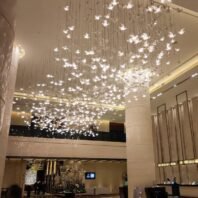 An awe-inspiring custom lighting installation dominates the ceiling of a luxurious hotel lobby. The chandelier features hundreds of illuminated butterfly-shaped elements suspended at various heights, creating a mesmerizing, dream-like effect. Each delicate butterfly glows softly, emitting a warm, diffused light that bathes the space in a gentle radiance. The installation spans the entire ceiling, giving the illusion of a vast swarm of luminous butterflies frozen in mid-flight. This enchanting lighting design is complemented by the lobby's modern architecture, including tall cylindrical columns and a visible mezzanine level. The reception desk is visible in the foreground, while decorative elements and a digital display can be seen in the background. The combination of the whimsical, nature-inspired lighting with the sleek, contemporary interior transforms the lobby into a captivating and welcoming space that immediately impresses hotel guests upon entry.