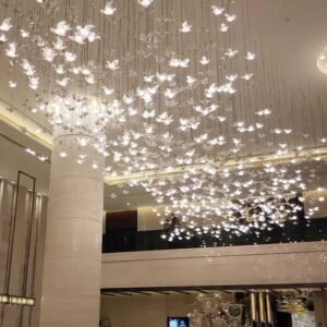 A breathtaking custom lighting installation adorns the ceiling of a spacious hotel lobby. The chandelier features hundreds of illuminated butterfly-shaped elements suspended at varying heights, creating an enchanting, ethereal effect. Each delicate butterfly glows softly, casting a warm, diffused light throughout the space. The installation spans a large area of the ceiling, giving the impression of a swarm of luminous butterflies in mid-flight. This magical lighting design is complemented by the lobby's modern architecture, including tall columns and a mezzanine level visible in the background. The combination of the playful, nature-inspired lighting with the sleek, contemporary interior transforms the lobby into a captivating and welcoming space for hotel guests.