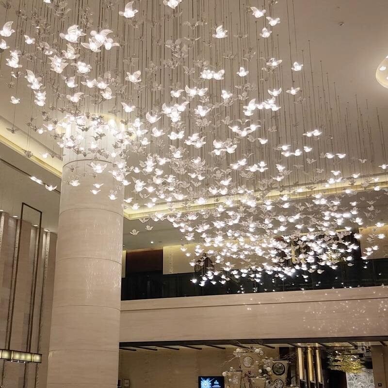 A breathtaking custom lighting installation adorns the ceiling of a spacious hotel lobby. The chandelier features hundreds of illuminated butterfly-shaped elements suspended at varying heights, creating an enchanting, ethereal effect. Each delicate butterfly glows softly, casting a warm, diffused light throughout the space. The installation spans a large area of the ceiling, giving the impression of a swarm of luminous butterflies in mid-flight. This magical lighting design is complemented by the lobby's modern architecture, including tall columns and a mezzanine level visible in the background. The combination of the playful, nature-inspired lighting with the sleek, contemporary interior transforms the lobby into a captivating and welcoming space for hotel guests.