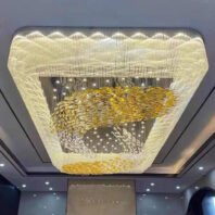 A magnificent ceiling-mounted chandelier showcasing opulent custom lighting design for luxury hotels. The chandelier features a large hexagonal frame with intricate crystal and glass elements creating a mesmerizing display. The outer edge of the frame is adorned with a wave-like pattern of small lights, giving the impression of a shimmering border. Inside the frame, golden yellow clusters swirl in dynamic patterns, reminiscent of abstract clouds or celestial bodies. These are interspersed with silver droplets and hanging crystal strands, adding depth and sparkle to the design. The chandelier is suspended from a recessed ceiling with additional ambient lighting. This grand lighting fixture transforms the space, likely a hotel atrium or grand lobby, into a dazzling, light-filled environment that exudes luxury and sophistication.