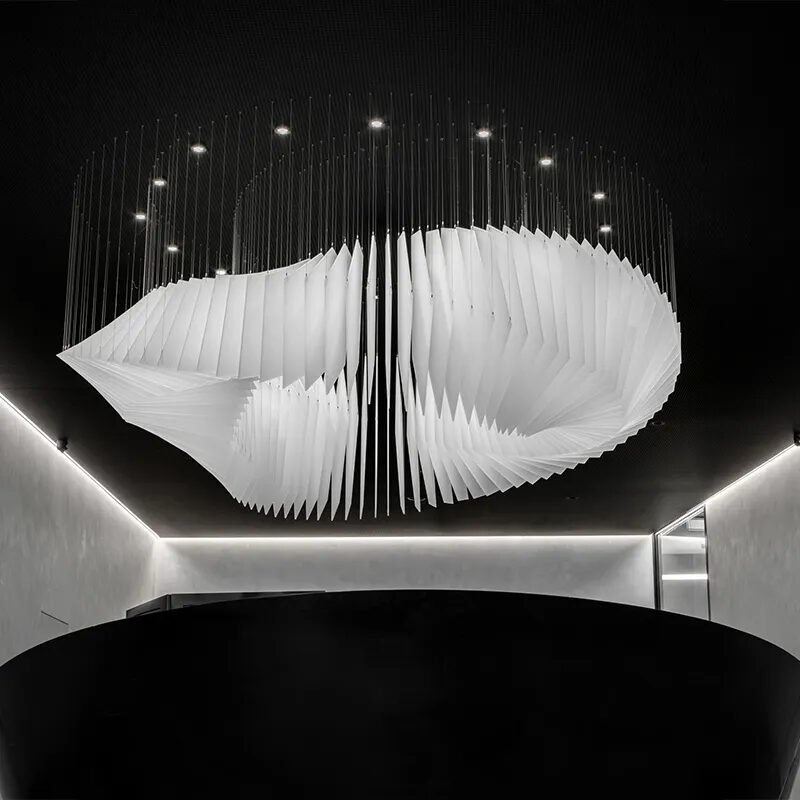 A breathtaking large chandelier showcasing innovative custom lighting design for luxury hotels. This big chandelier features a series of white, angular panels arranged in a fluid, wing-like formation, suspended from thin wires. The hotel chandelier's unique composition creates a striking interplay of light and shadow, with each panel precisely angled to reflect and diffuse light. Floating above a sleek, curved black reception desk, the chandelier becomes the focal point of the minimalist space. Small spotlights above accentuate its sculptural quality, while recessed lighting around the ceiling perimeter adds depth. This avant-garde custom lighting installation transforms the modern lobby into an artistic display, perfect for creating a sophisticated atmosphere in high-end hospitality environments such as boutique hotel entrances or upscale art galleries.