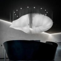 A striking large chandelier showcasing innovative custom lighting design for luxury hotels. This big chandelier features an undulating white fabric structure suspended from thin metal wires, creating a cloud-like ethereal effect. The hotel chandelier's unique composition resembles a billowing sail or abstract wave, with gentle curves and sharp angles. Suspended above a sleek black curved reception desk in a minimalist space, the chandelier becomes a captivating focal point. Strategically placed LED lights within the structure cast a soft, diffused glow, while recessed ceiling lights add depth. This avant-garde custom lighting installation transforms the contemporary space into a dramatic, sculptural display, perfect for creating a sophisticated atmosphere in high-end hospitality environments such as boutique hotel lobbies or modern art galleries.