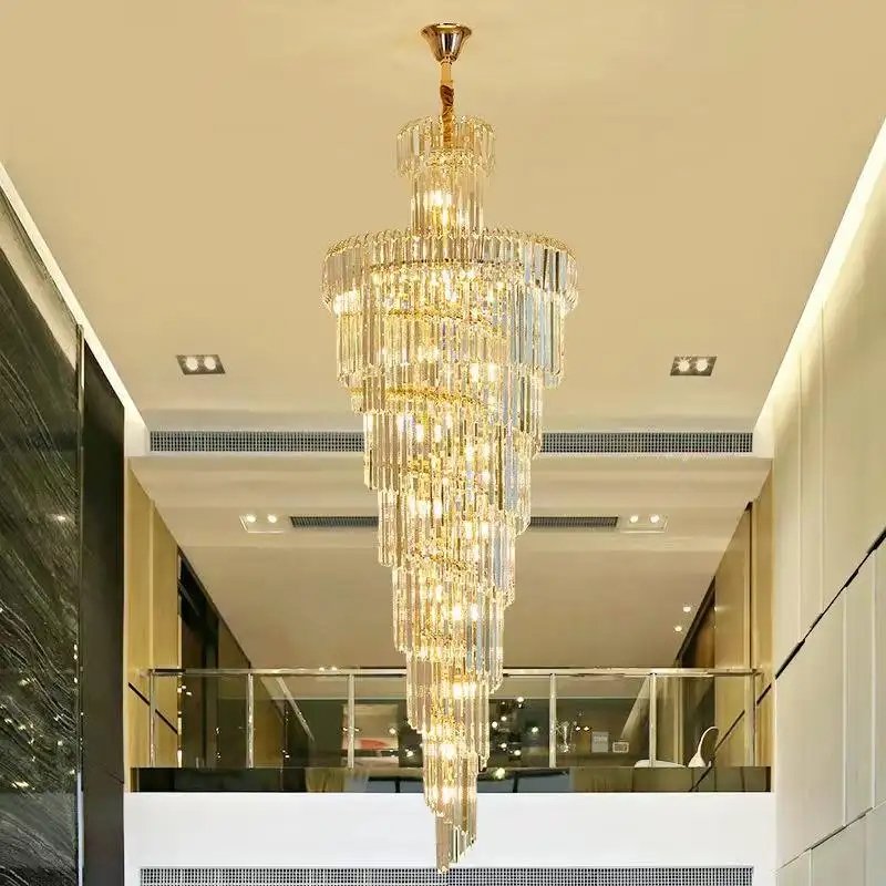 A stunning large chandelier showcasing exquisite custom lighting design for luxury hotels. This big chandelier features multiple tiers of crystal prisms arranged in a spiraling, tapered shape, creating a dazzling waterfall effect. The hotel chandelier spans two floors, suspended from a gold-accented ceiling fixture. Positioned in a modern atrium with cream-colored walls and sleek glass balustrades, the chandelier becomes a captivating focal point. Its crystal elements reflect warm light, illuminating the contemporary space with a golden glow. The chandelier's elegant design contrasts beautifully with the minimalist surroundings, including recessed lighting and a mezzanine level visible in the background. This breathtaking custom lighting installation transforms the space into a luxurious, light-filled environment, perfect for creating a sophisticated atmosphere in high-end hospitality settings such as upscale hotel lobbies or grand entrances.