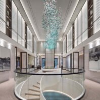 A stunning large chandelier showcasing innovative custom lighting design for luxury hotels. This big chandelier features thousands of suspended glass elements in varying shades of blue and teal, creating a mesmerizing waterfall effect. The hotel chandelier's unique composition resembles a cascading stream of water droplets, starting sparsely at the top and densifying towards the bottom. Suspended in a towering modern atrium with sleek white walls and perforated metal panels, the chandelier becomes a breathtaking focal point. The space features a circular glass balustrade on the ground floor, leading to a sunken area. Ambient lighting accentuates the chandelier's azure hues, while artwork adorns the walls. This exquisite custom lighting installation transforms the contemporary space into a captivating, aquatic-inspired display, perfect for creating a dramatic atmosphere in high-end hospitality environments such as grand lobbies or upscale hotels.