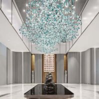 A breathtaking large chandelier showcasing innovative custom lighting design for luxury hotels. This big chandelier features thousands of suspended glass elements in shades of clear, light blue, and teal, creating a mesmerizing cloud-like effect. The hotel chandelier's unique composition resembles a sparkling nebula, with densely clustered glass pieces that gradually disperse towards the edges. Suspended in a high-ceilinged modern lobby with sleek white walls and vertical gray panels, the chandelier becomes a stunning focal point. Below, a rectangular black reflective pool mirrors the chandelier, doubling its visual impact. Flanking the pool are two tall sculptures, while a striking red-dotted artwork adorns the far wall. This exquisite custom lighting installation transforms the contemporary space into a captivating, celestial-inspired display, perfect for creating a dramatic and sophisticated atmosphere in high-end hospitality environments such as grand entrances or upscale hotel lobbies.