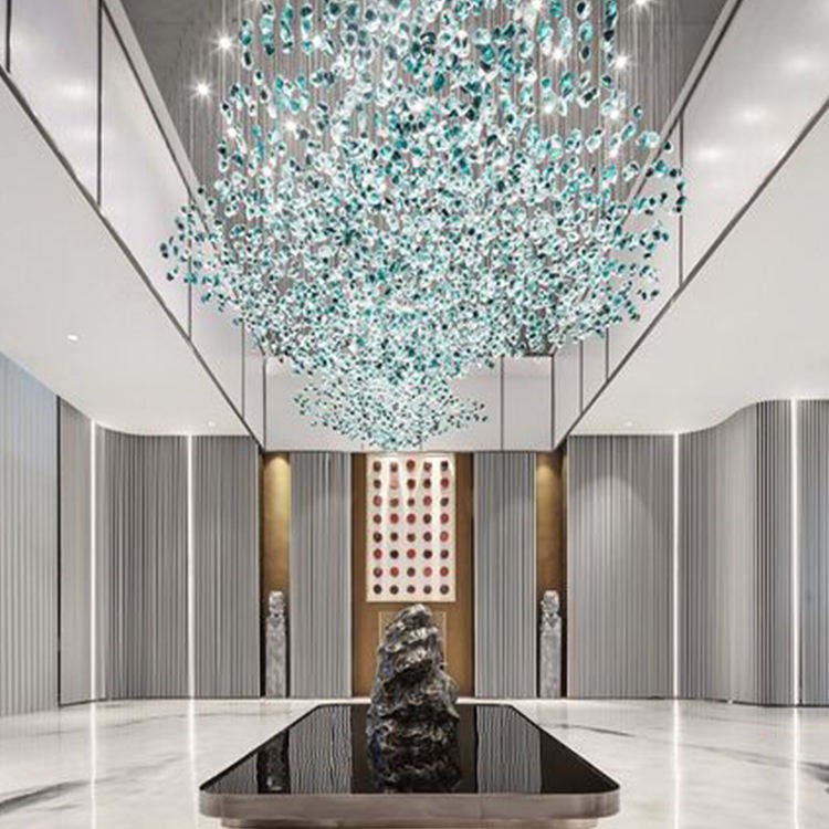 A breathtaking large chandelier showcasing innovative custom lighting design for luxury hotels. This big chandelier features thousands of suspended glass elements in shades of clear, light blue, and teal, creating a mesmerizing cloud-like effect. The hotel chandelier's unique composition resembles a sparkling nebula, with densely clustered glass pieces that gradually disperse towards the edges. Suspended in a high-ceilinged modern lobby with sleek white walls and vertical gray panels, the chandelier becomes a stunning focal point. Below, a rectangular black reflective pool mirrors the chandelier, doubling its visual impact. Flanking the pool are two tall sculptures, while a striking red-dotted artwork adorns the far wall. This exquisite custom lighting installation transforms the contemporary space into a captivating, celestial-inspired display, perfect for creating a dramatic and sophisticated atmosphere in high-end hospitality environments such as grand entrances or upscale hotel lobbies.