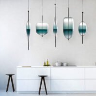 A sleek large chandelier showcasing innovative custom lighting design for contemporary hotels. This big chandelier features five suspended glass elements in varying shapes - elongated ovals, rounded squares, and circles. Each piece displays a striking gradient from clear to teal, resembling elegant vessels filled with water. The hotel chandelier hangs above a pristine white kitchen island, creating a floating, ethereal effect. This exquisite custom lighting installation transforms the minimalist space into a sophisticated display, perfect for upscale hospitality environments. The chandelier's unique composition adds a touch of artistic flair and serene ambiance to the modern kitchen setting.