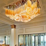 A magnificent large chandelier showcasing innovative custom lighting design for luxury hotels. This big chandelier features thousands of amber-colored glass tubes arranged in undulating waves, creating a mesmerizing golden cascade effect. The hotel chandelier's unique composition is complemented by twinkling lights above, forming intricate swirling patterns. Suspended from an ornate coffered ceiling in a grand lobby, the chandelier becomes a stunning focal point. Classical columns with gilded Corinthian capitals and decorative wall moldings frame the space, while large glass doors offer a view of the exterior. This breathtaking custom lighting installation transforms the elegant space into a warm, light-filled environment, perfect for creating a luxurious atmosphere in high-end hospitality settings such as prestigious hotel entrances or upscale reception areas.