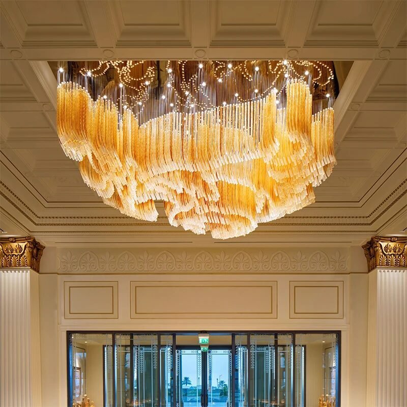 A spectacular large chandelier showcasing exquisite custom lighting design for luxury hotels. This big chandelier features thousands of golden glass tubes cascading in undulating waves, creating a mesmerizing waterfall effect. The hotel chandelier's unique composition is accentuated by twinkling lights above, forming intricate swirling patterns. Suspended from an ornate coffered ceiling in a grand entryway, the chandelier becomes a stunning focal point. Classical columns with gilded Corinthian capitals frame the space, while large glass doors offer a glimpse of the exterior. This breathtaking custom lighting installation transforms the elegant space into a dazzling, light-filled environment, perfect for creating a luxurious atmosphere in high-end hospitality settings such as prestigious hotel lobbies or grand ballroom entrances.