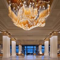 A breathtaking large chandelier showcasing exquisite custom lighting design for luxury hotels. This big chandelier features a stunning array of golden glass tubes and twinkling lights, forming an intricate floral pattern that spans the ceiling. The hotel chandelier's unique composition resembles a blooming golden flower, with delicate tendrils of light creating swirling patterns above. Suspended in a grand lobby with classical columns adorned with gold Corinthian capitals, the chandelier becomes the focal point of the space. The room's marble floors reflect the warm glow, while large windows offer a view of the twilight sky and palm trees outside. This magnificent custom lighting installation transforms the opulent space into a dazzling, light-filled environment, perfect for creating a luxurious atmosphere in high-end hospitality settings such as prestigious hotel entrances.