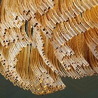 A close-up view of an extraordinary large chandelier showcasing innovative custom lighting design for luxury hotels. This big chandelier features hundreds of curved, translucent glass tubes in golden hues, creating a mesmerizing waterfall effect. The hotel chandelier's unique composition resembles a swirling, abstract sunburst with each glass element carefully arranged to form fluid, organic patterns. Warm light filters through the amber-tinted glass, casting a soft, golden glow. This exquisite custom lighting installation transforms any space into a striking, artistic display, perfect for creating a sophisticated and modern atmosphere in high-end hospitality environments such as upscale hotel lobbies, grand ballrooms, or contemporary art-inspired venues.