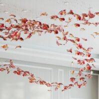 A breathtaking large chandelier showcasing innovative custom lighting design for upscale hotels. This big chandelier features hundreds of suspended glass elements shaped like abstract autumn leaves in vibrant red, orange, and golden hues. The hotel chandelier's unique composition creates a sweeping, dynamic effect, resembling a gust of wind carrying fallen leaves across the ceiling. Each leaf-like piece is intricately crafted with swirling patterns and suspended on nearly invisible wires, giving the illusion of floating. Recessed ceiling lights add depth and sparkle to the installation. This exquisite custom lighting transforms the white, minimalist space into an enchanting, nature-inspired display, perfect for creating a dramatic and sophisticated atmosphere in high-end hospitality environments such as grand lobbies or ballrooms.