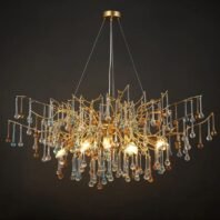 An opulent chandelier featuring an intricate gold-toned branch structure adorned with numerous crystal droplets in various colors. The fixture has an organic, tree-like design with golden branches extending outward and downward in a graceful, asymmetrical pattern. Hanging from these branches are crystal pendants in clear, light blue, and amber hues, creating a multi-colored sparkling effect. The chandelier is illuminated from within, with visible warm light bulbs nestled among the golden branches, casting a soft glow through the crystals. The fixture is suspended from a circular gold ceiling mount by thin wires. Photographed against a dark background, the chandelier's complex design and the interplay between the metallic gold and colorful crystals are highlighted, showcasing its blend of modern artistry and classic elegance.