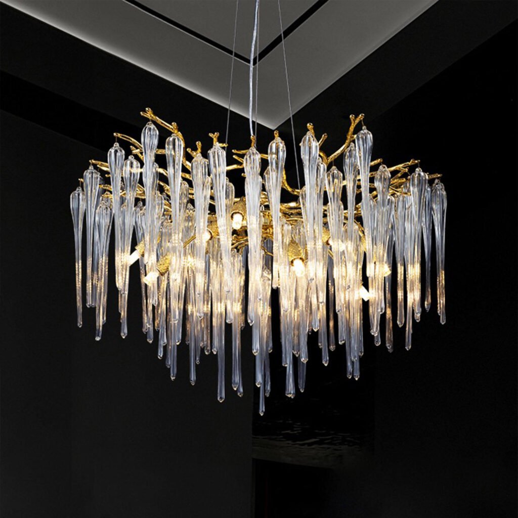 An opulent chandelier featuring numerous crystal icicle-like pendants suspended from gold-toned branches. The chandelier has a circular shape with crystal elements of varying lengths creating a cascading effect. The gold branches form an intricate, organic structure resembling tree limbs or coral. The fixture is illuminated from within, causing the crystal pendants to glow warmly and creating a striking contrast against the dark background. The chandelier is suspended by thin wires from a ceiling with recessed lighting, emphasizing its floating appearance. The overall design combines natural, organic forms with luxurious materials, resulting in a stunning centerpiece that blends modern and classic aesthetics.