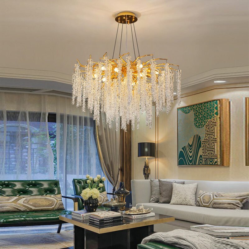 An opulent living room featuring a stunning crystal and gold chandelier as the centerpiece. The chandelier has cascading crystal strands suspended from gold-toned branches, creating a waterfall effect. The room is decorated in a modern Art Deco style with a cream-colored sofa, a gold and black coffee table, and emerald green accents. A large window with sheer curtains allows natural light in, while a bold geometric artwork in green and gold adorns the wall. The space is furnished with various decorative elements including flowers, books, and a floor lamp, creating a harmonious blend of luxury and comfort.