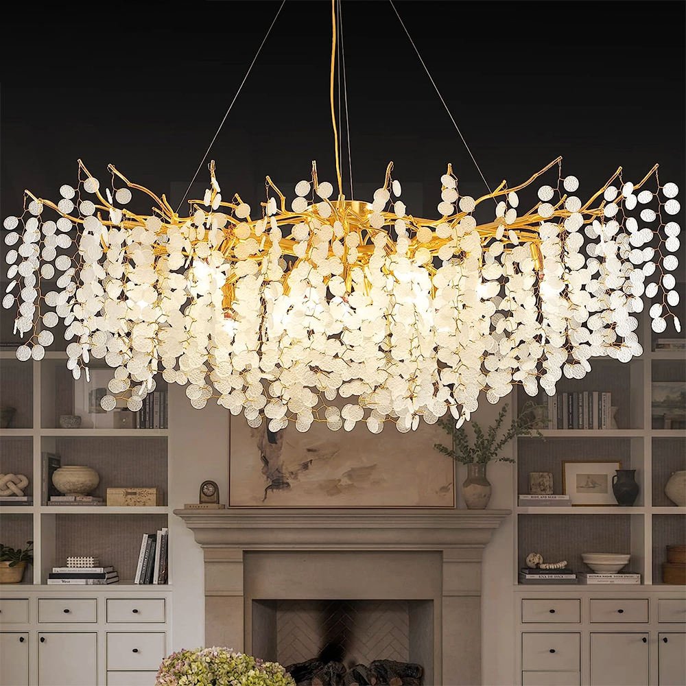 An opulent oval-shaped crystal chandelier hangs in a sophisticated living room. The chandelier features golden branches adorned with numerous cascading crystal discs, creating a waterfall effect. It's illuminated, casting a warm glow over the space. The chandelier is suspended above a classic fireplace flanked by built-in bookshelves. The room's decor includes neutral tones, with the shelves displaying books, vases, and decorative objects. A painting is visible above the fireplace mantel, and flowers are placed in the foreground, adding a touch of natural beauty to the refined interior.