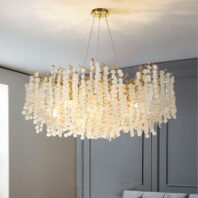 A stunning oval-shaped chandelier featuring a multitude of crystal discs hanging from gold-toned branches. The chandelier is suspended from a gold circular base on the ceiling by thin wires. It emits a warm, golden glow, illuminating the textured crystal elements that create a cascading, waterfall-like effect. The fixture is set against a backdrop of light gray walls with modern paneling, blending contemporary and classic design elements. The chandelier's intricate design creates a striking contrast with the room's clean lines and muted color palette. A portion of what appears to be a light-colored sofa or furniture piece is visible at the bottom of the image, suggesting a sophisticated living space. The overall effect is one of modern luxury with a touch of traditional elegance.