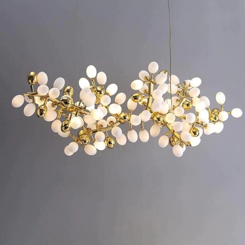 A contemporary horizontal chandelier featuring a cluster of white frosted glass orbs and golden accents. The fixture consists of numerous egg-shaped white glass globes arranged in an organic, branch-like formation. Interspersed among the white orbs are smaller, highly reflective golden spheres, adding a luxurious contrast. The structure supporting these elements is made of thin, gold-toned metal arms that branch out in various directions, creating a complex and intricate design. The overall shape of the chandelier is elongated and asymmetrical, resembling a stylized molecular structure or a modern interpretation of a flowering branch. Several of the white orbs are illuminated, emitting a soft, warm glow. The chandelier is suspended by thin wires against a neutral grey background, highlighting its intricate design and the interplay between the matte white glass and shiny gold elements.