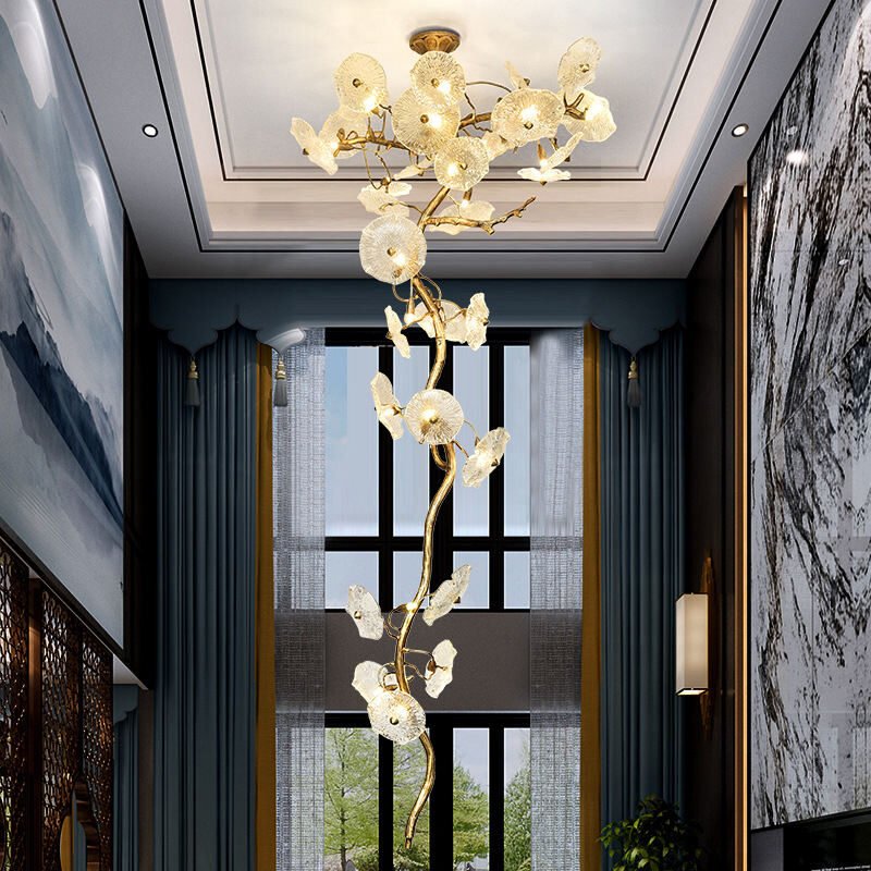 A stunning, vertically elongated chandelier featuring glass flower-like elements on a golden branch, spanning a luxurious two-story interior space. The chandelier resembles a tall, artistic tree with illuminated glass blooms. Its golden stem twists upwards, adorned with numerous translucent, textured glass shades that resemble flowers or sea anemones. Each glass element houses a light, creating a warm, ethereal glow. The fixture is suspended from a white coffered ceiling and extends down through the room, framed by floor-to-ceiling windows with dark blue curtains and sheer drapes. The space features dark walls, contrasting with the bright chandelier. Large abstract artworks in black and white tones adorn the walls, adding to the sophisticated ambiance. The room's design combines modern luxury with organic, nature-inspired elements, creating a striking and elegant atmosphere.