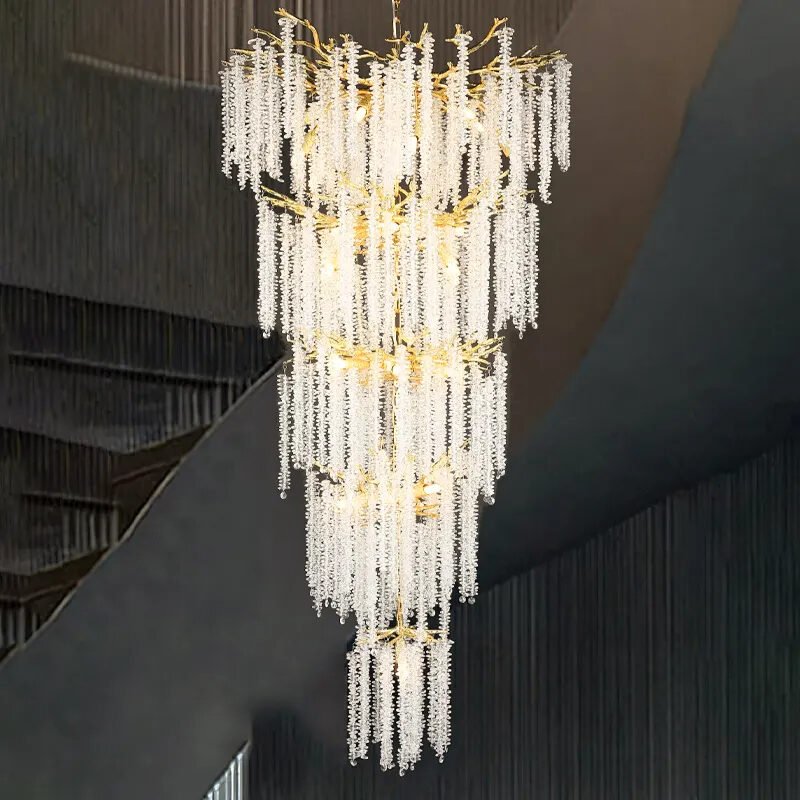 Grand multi-tiered crystal chandelier with cascading strands and gold accents. Illuminated from within, creating a warm glow. Hangs in a modern space with curved architectural elements. Chandelier's sparkle contrasts with dark surroundings.