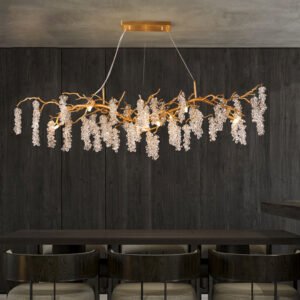 A modern dining room featuring a striking horizontal chandelier that resembles golden branches adorned with cascading crystal clusters. The chandelier hangs from a rectangular gold ceiling mount above a dark wooden dining table. Three chairs with curved backs are partially visible beneath the table. The room has a dark, textured wood-paneled wall, creating a dramatic backdrop for the illuminated chandelier. The contrast between the warm golden tones of the chandelier's branches and the sparkling crystals against the dark surroundings creates a luxurious and intimate atmosphere in the dining space.