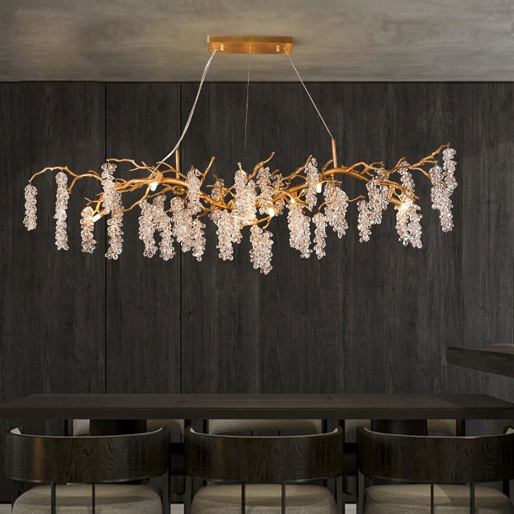 A modern dining room featuring a striking horizontal chandelier that resembles golden branches adorned with cascading crystal clusters. The chandelier hangs from a rectangular gold ceiling mount above a dark wooden dining table. Three chairs with curved backs are partially visible beneath the table. The room has a dark, textured wood-paneled wall, creating a dramatic backdrop for the illuminated chandelier. The contrast between the warm golden tones of the chandelier's branches and the sparkling crystals against the dark surroundings creates a luxurious and intimate atmosphere in the dining space.