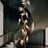 A luxurious interior featuring a dramatic spiral staircase with sleek, curved black railings. Alongside the staircase hangs a stunning vertical chandelier designed as a golden twisted branch with multiple illuminated glass bird figures. The birds appear to be in flight, creating a sense of movement. The chandelier extends from floor to ceiling, complementing the height of the staircase. The background has a deep, rich green color, providing a elegant contrast to the golden chandelier and the modern architecture of the staircase. This design beautifully merges contemporary architectural elements with nature-inspired artistic lighting.